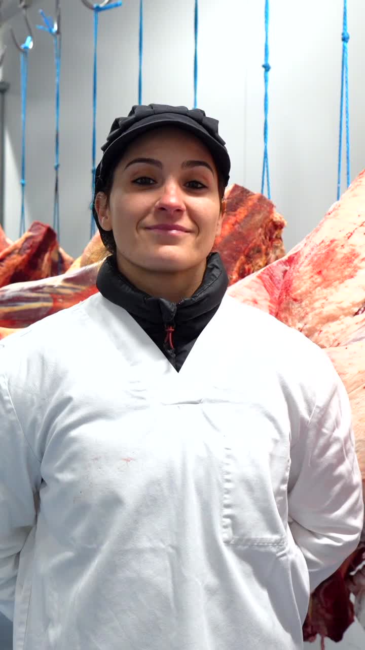 A woman in a white coat and hat stands in front of hanging meat