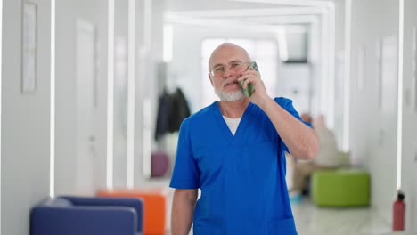 A-confident-elderly-man-with-a-beard-a-doctor-walks-along-the-corridor-of-a-bright-clinic-and-talks-on-the-phone-during-the-working-day.-A-confident-elderly-doctor-walks-through-the-clinic-talking