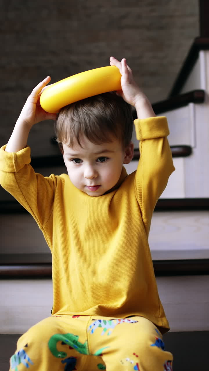 Funny toddler boy sitting on the stairs holding the ring from pyramid on his head. Kid playing at home with toys. Vertical video