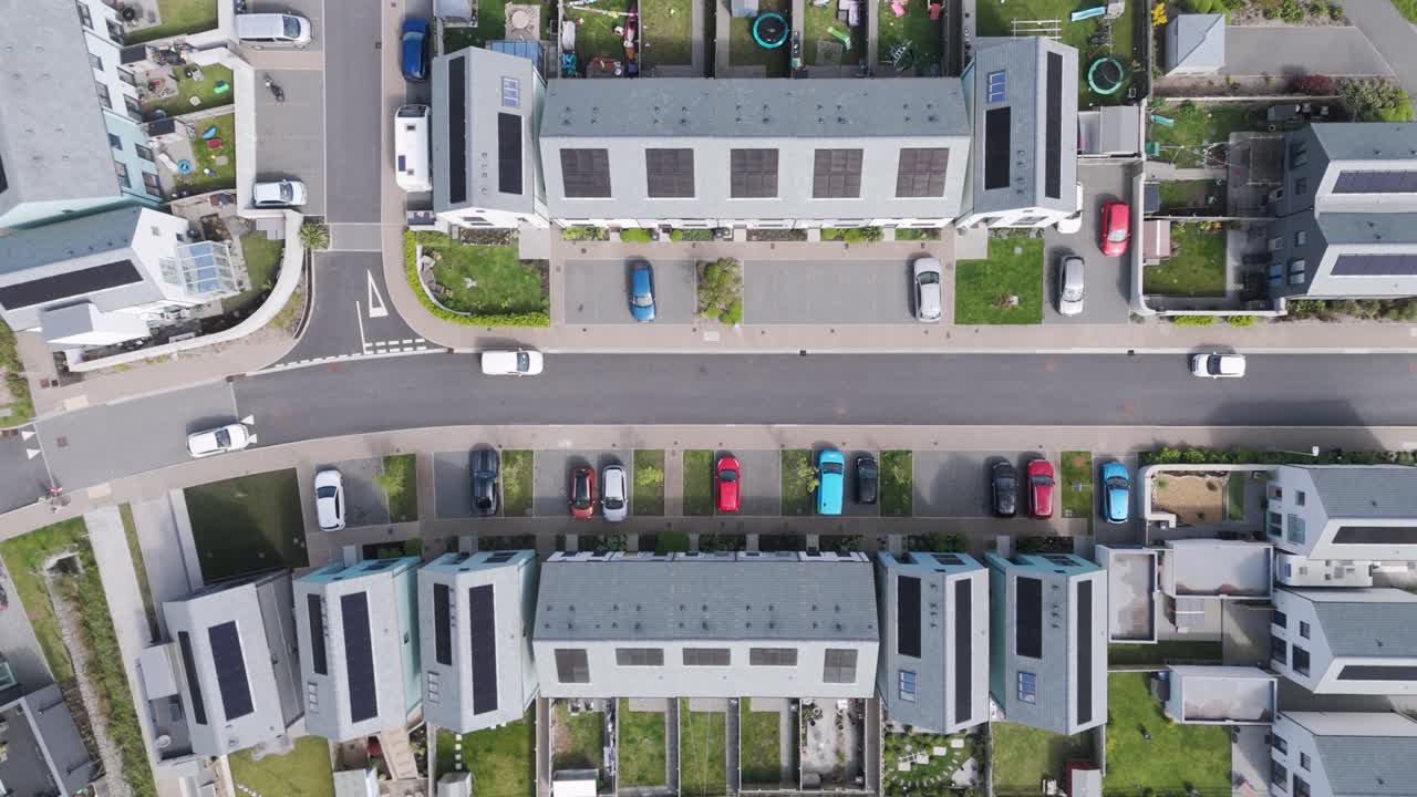 Top-down pull-out aerial of neatly curving suburban street lined by boxy contemporary homes with dark solar rooftops, driveways, and landscaped verges conveying sustainable living