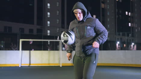 futbolista con capucha joggling fútbol durante la práctica nocturna en el campo deportivo urbano rodeado de edificios de gran altura y luces de calle brillantes