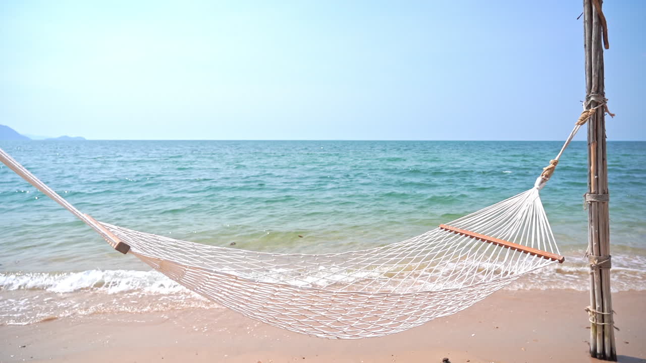 열대 섬의 청록색 바다 앞 해변의 모래에 박힌 두 개의 대나무 기둥 사이에 스프레더 바가 있는 빈 흰색 코톰 로프 그물 해목
