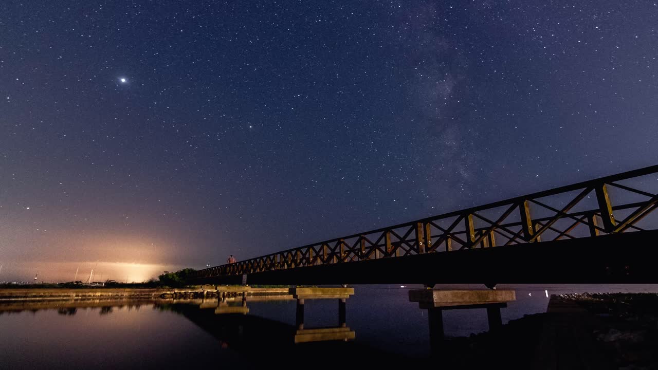 A mesmerizing timelapse of the Milky Way in Grado, Italy, with a scenic bridge adding depth to the cosmic view. A stunning blend of nature and structure under the night sky.
