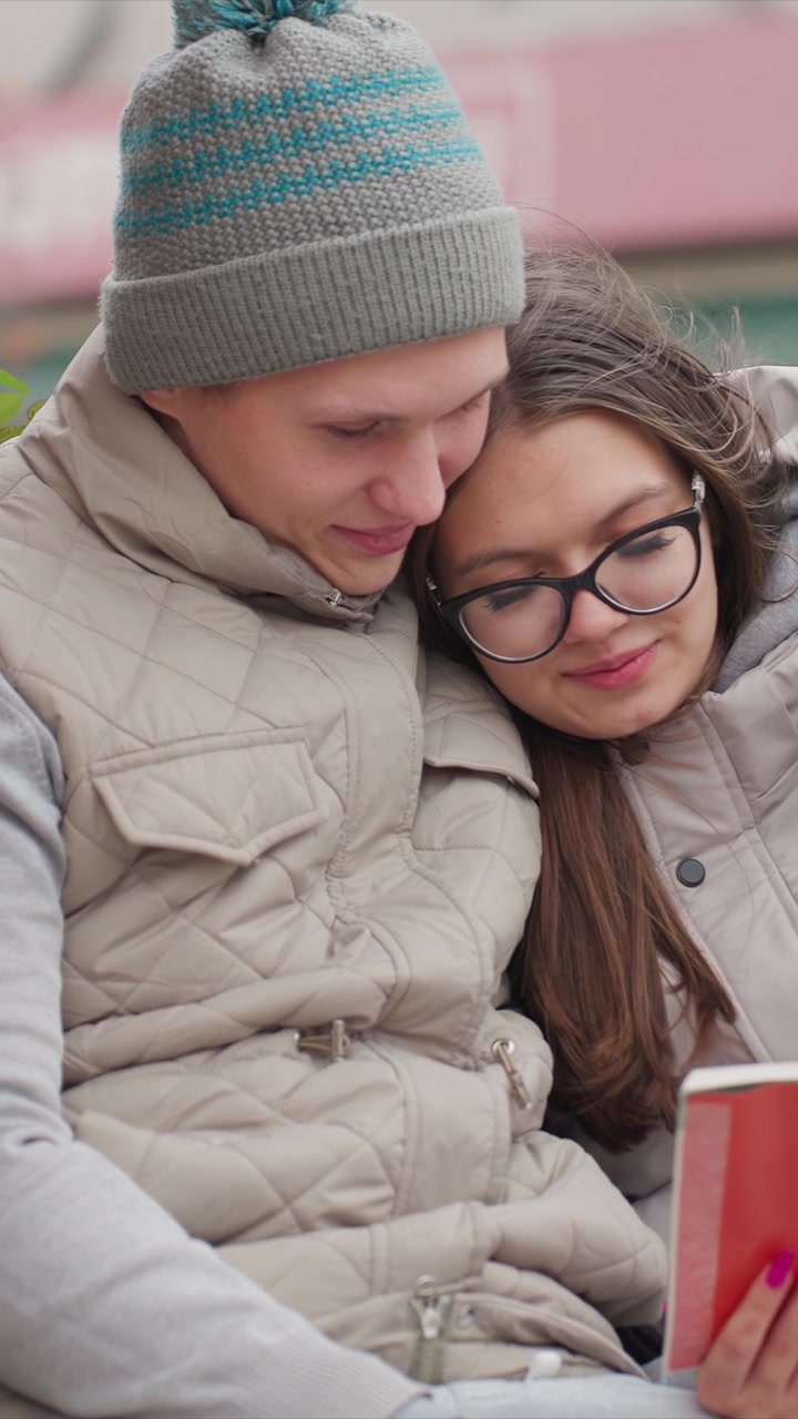 Individui immobili l'uno accanto all'altro che esaminano. Una donna appoggia la testa mentre tiene in mano un volume rosso aperto. L'ambientazione è un luogo all'aperto, probabilmente un parco o una piazza pubblica