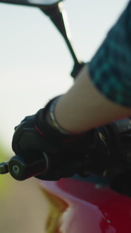 primer plano de la mano de un motociclista con guante y camisa a cuadros, acelerando una motocicleta, el fondo está borroso con un entorno verde natural