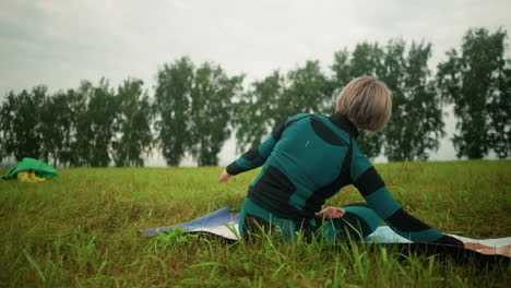 back view of woman lying on yoga mat practicing side bend pose in vast grassy field under cloudy skies, with yellow boots and green cloth in background, trees lining the horizon