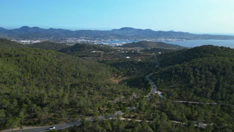 road winding through a vibrant green forest in ibiza, spain, leading down towards the beautiful san antonio bay. best aerial view flight