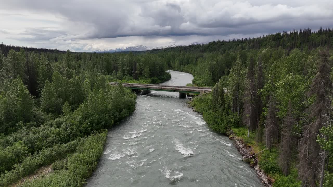 Silta joen yli metsässä, pilvinen taivas, lumihuippuiset vuoret kaukaisuudessa, Parks Highway -tie Alaskassa