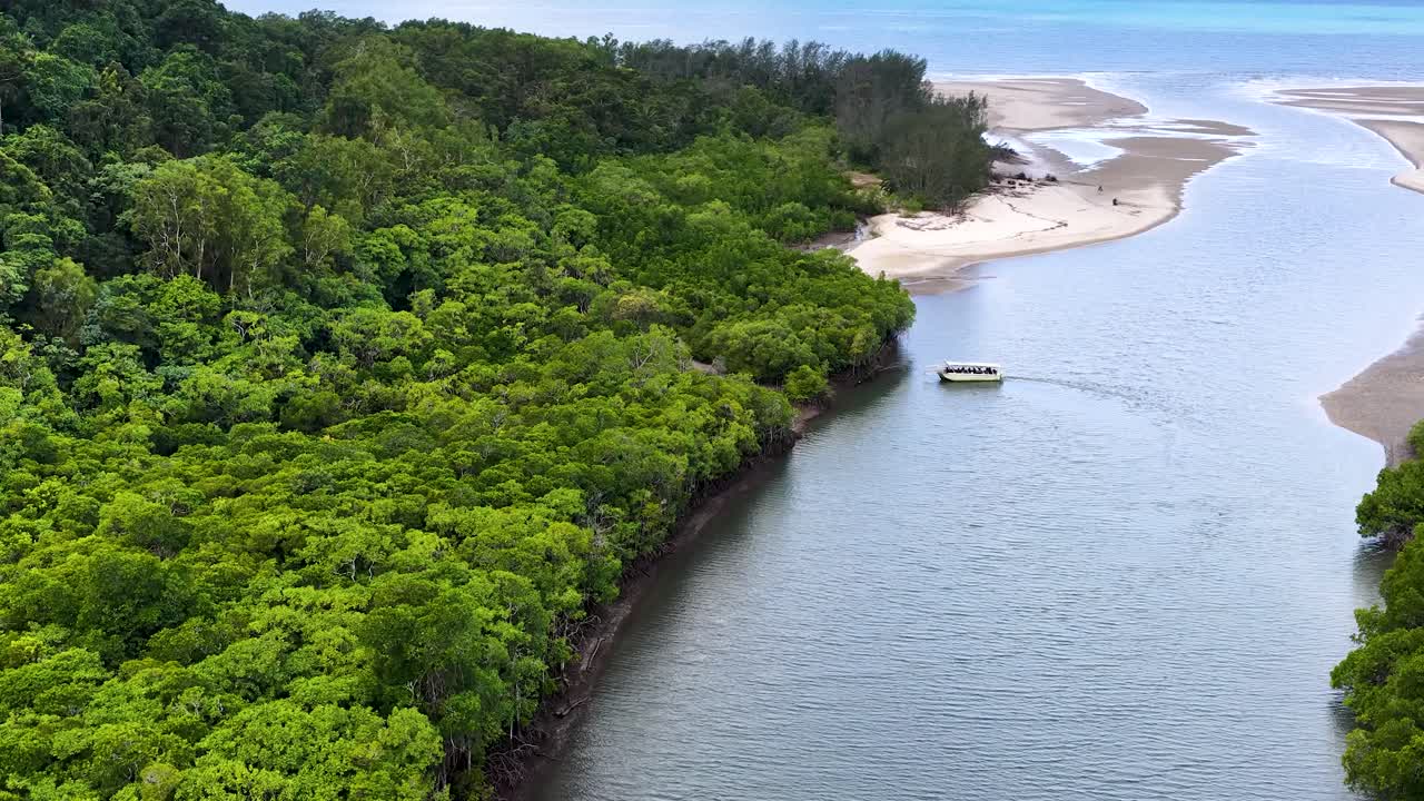 Drone tracks small boat navigating river beside lush mangroves, sandy beach, and rainforest canopy
