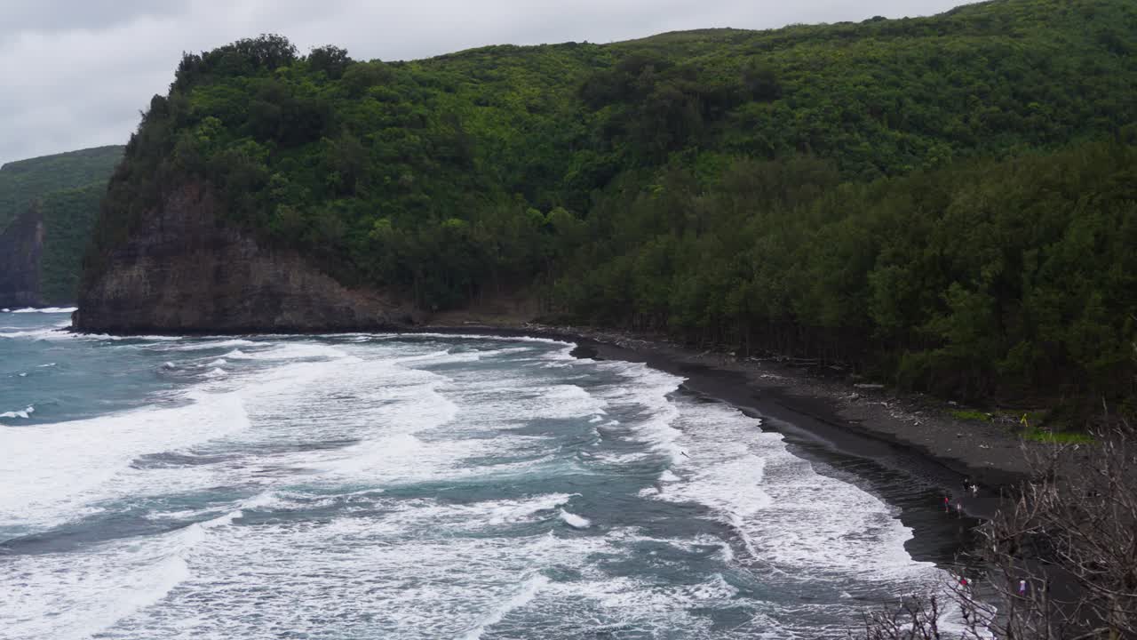 From a high lookout, white surf rakes a crescent of black sand beneath a dense green bluff, while gray water rolls ashore under soft overcast light and wind