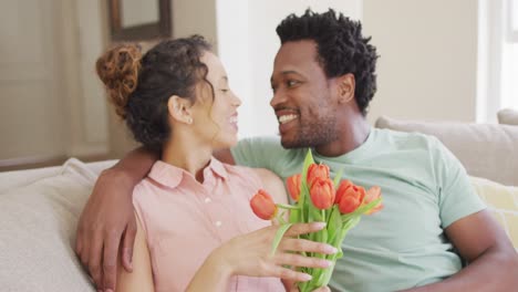 Happy-biracial-couple-sitting-on-sofa-with-bouquet-of-tulips