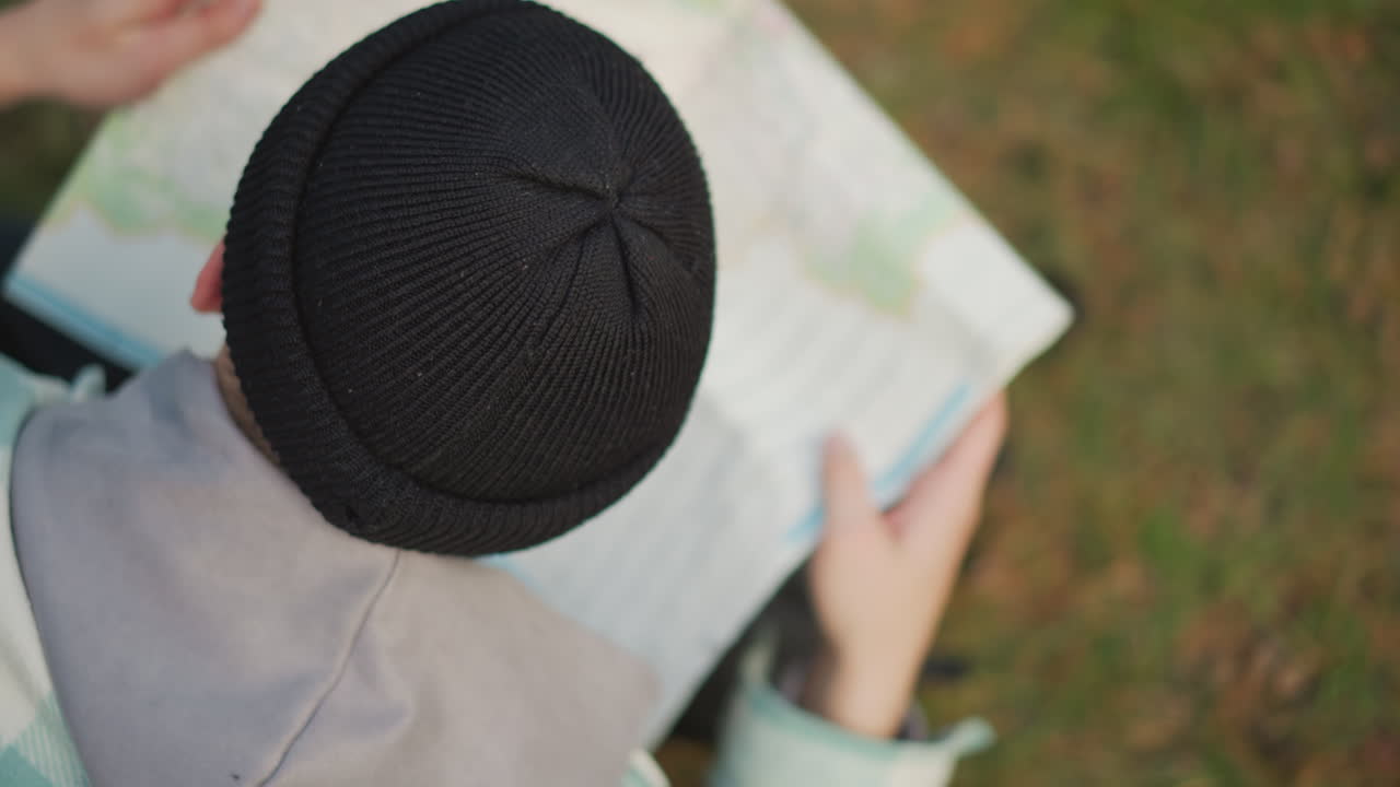 white male navigator scanning unfolded map overhead in woodland, selecting route and measuring distances, hands spread across paper, beanie and backpack at side, calm decision moment, textured folds