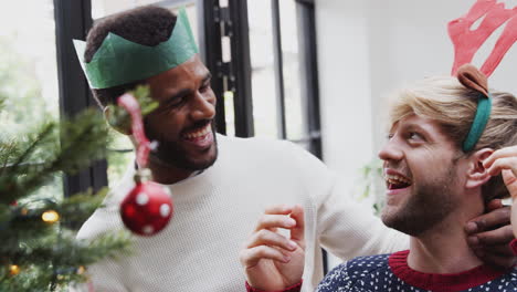 Mann-Setzt-Seinem-Männlichen-Partner-Ein-Kostümgeweih-Auf,-Während-Sie-Gemeinsam-Dekorationen-An-Den-Weihnachtsbaum-Hängen