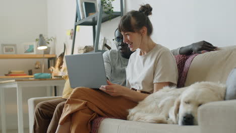 young couple sitting with dog on sofa and using laptop