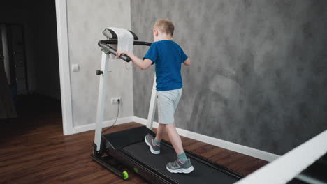 rear view of boy exercising on treadmill tapping machine handle with left hand while his shadow is clearly visible on gray wall in well-lit room with wooden floor and minimal decor