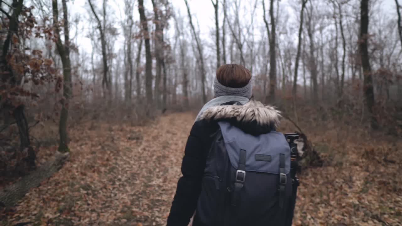 Following shot of female caucasian photographer with backpack and tripod walking on leaf covered dirt road in forest