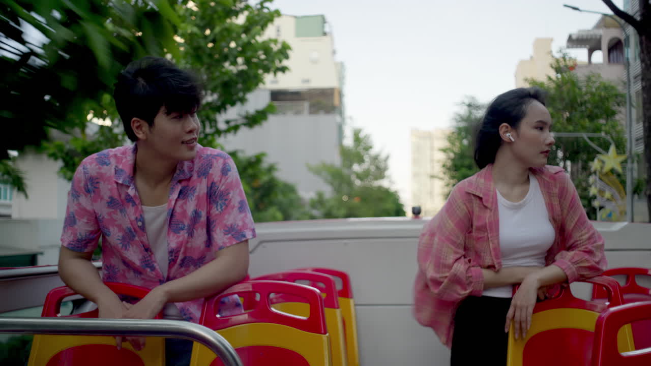 젊은 아시아 커플이 투어 버스에서 함께 여행하면서 현대 도시를 돌아다니며 헤드폰으로 음악을 듣고 있습니다.