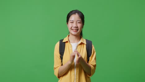 asian woman student with a backpack and some books smiling and clapping her hands while standing in the green screen background studio