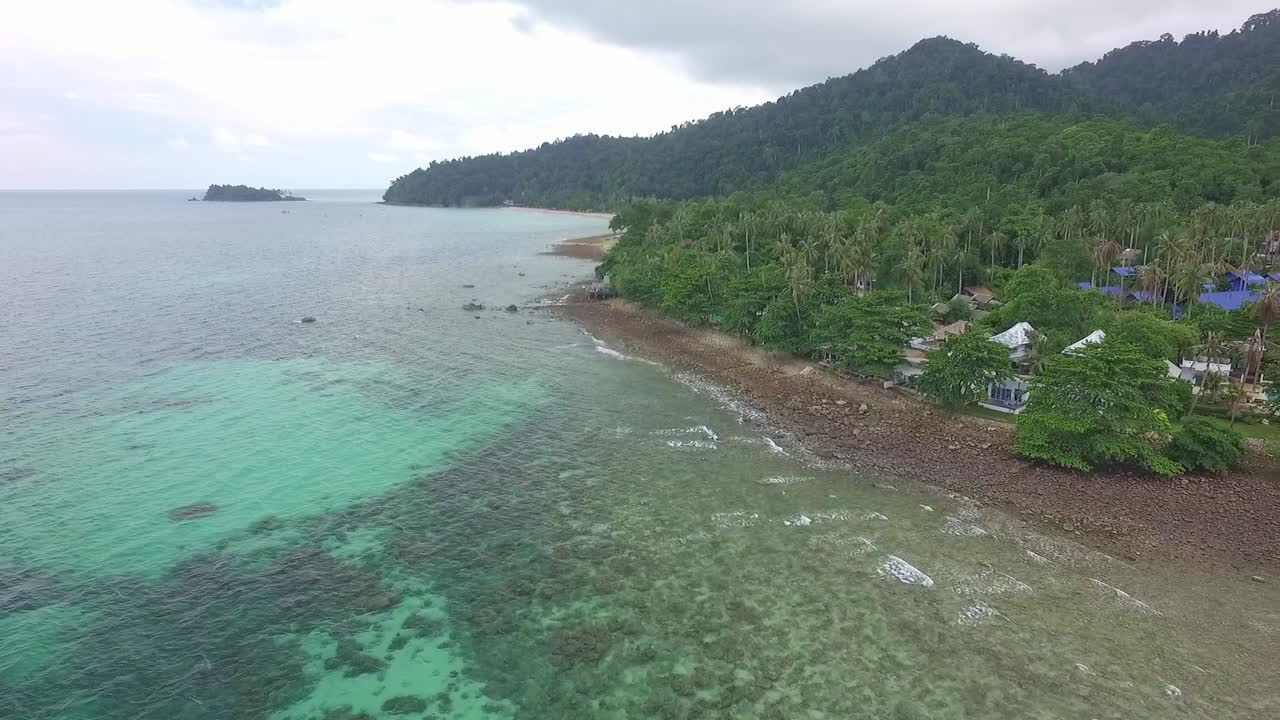 toma aérea de seguimiento de la costa de la isla tropical con océano, islas y sartenes alrededor para revelar pequeños resorts de estilo tailandés y torres de telecomunicaciones