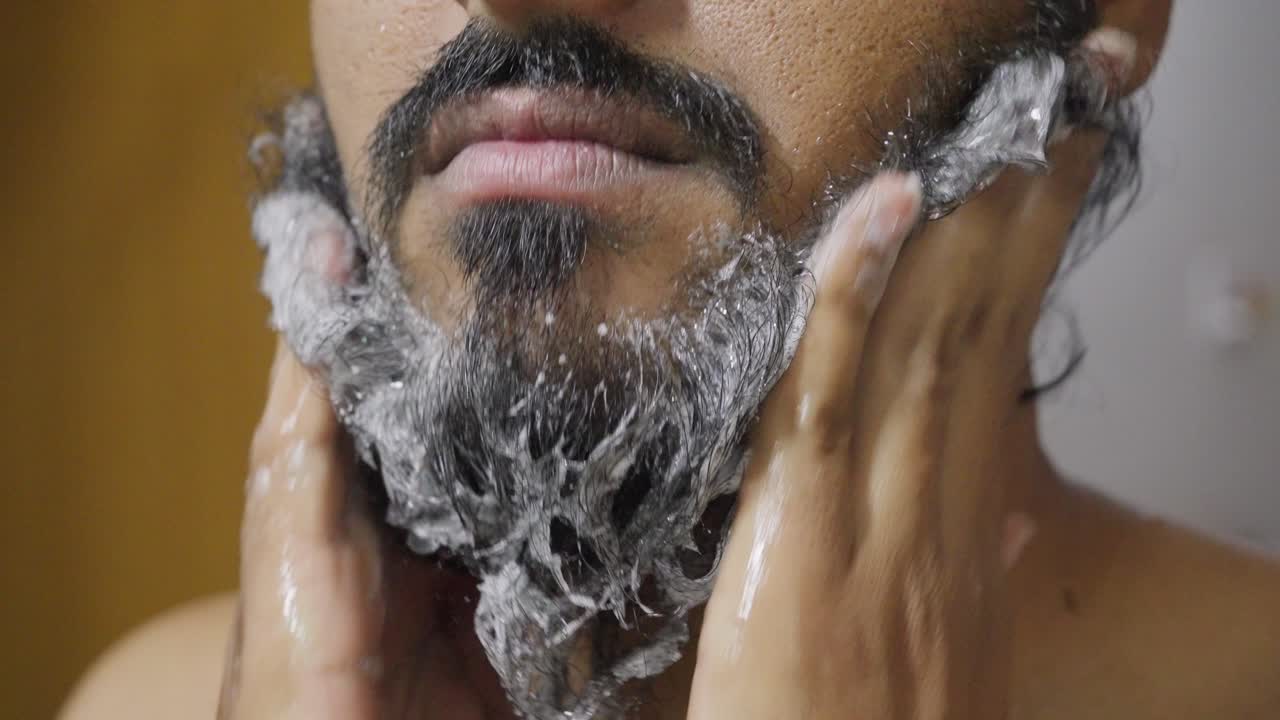 Primo piano di un uomo con la barba che la lava delicatamente con il sapone, concentrandosi sulle sue mani e sulla barba mentre la schiuma crea una texture pulita e morbida.