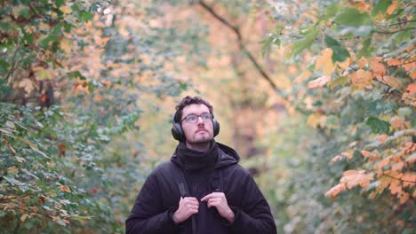 Young-European-man-with-headphones-in-a-vibrant-fall-mixed-forest,-observing-colorful-autumn-leaves