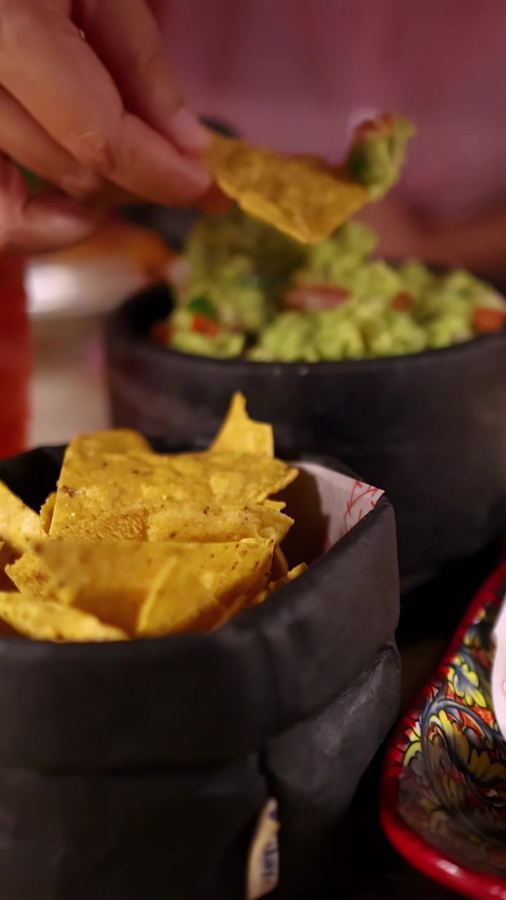A hand selects a nacho chip from a black basket and dips it into chunky guacamole, with salsa and a red cocktail in a warmly lit restaurant