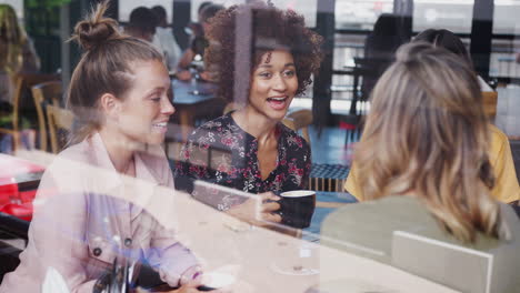 Four-Young-Female-Friends-Meeting-Sitting-At-Table-In-Coffee-Shop-And-Talking-Viewed-Through-Window