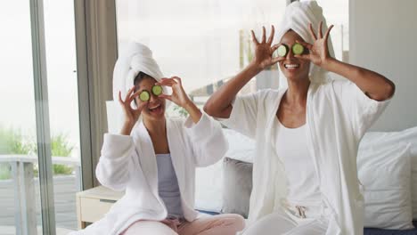 video of happy diverse female friends in robes having fun with cucumber slices