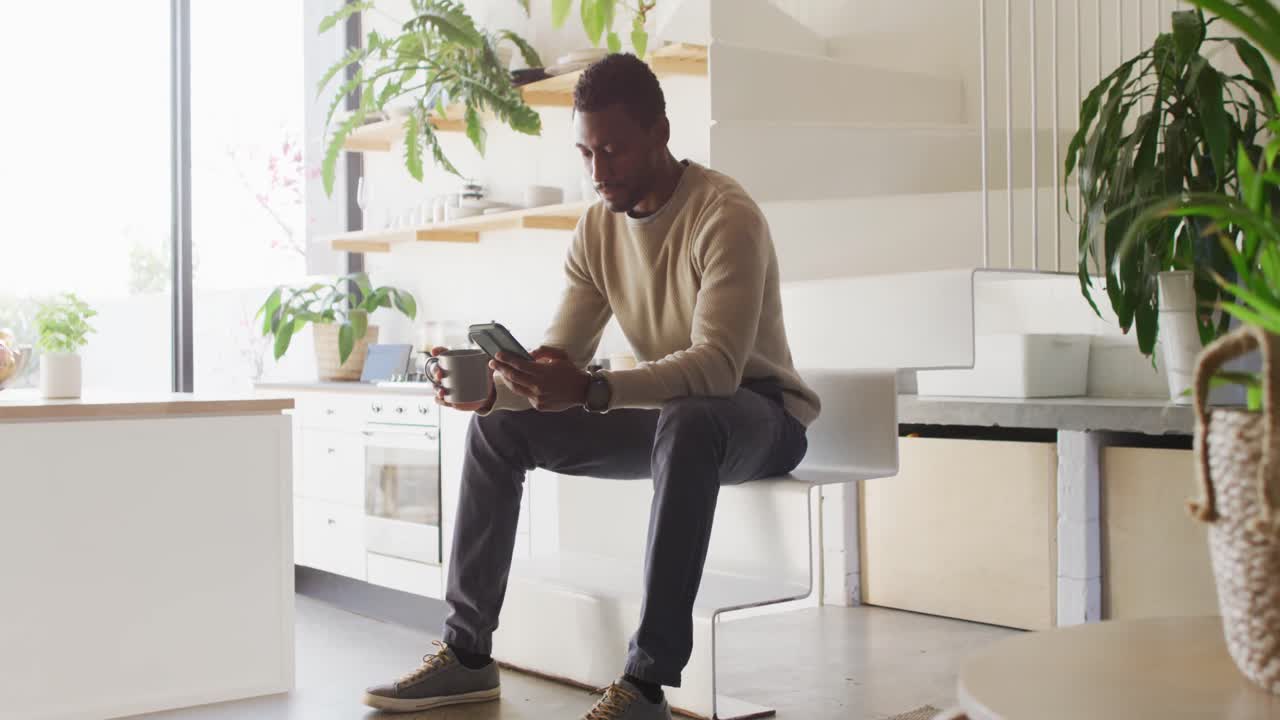 Happy african american man sitting on stairs in kitchen, drinking coffee and using smartphone