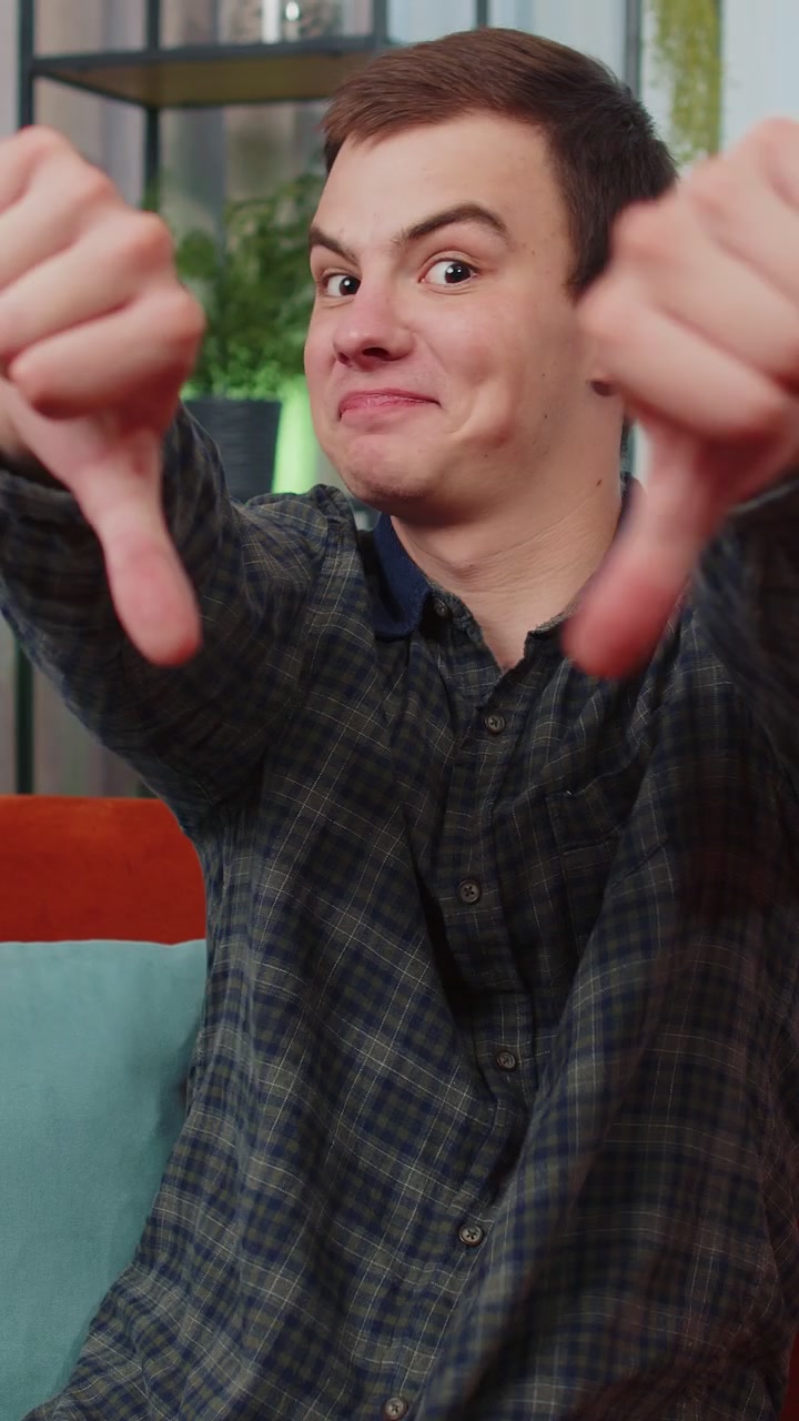 Upset young man showing thumbs down dislike bad work disapproval dissatisfied feedback at home