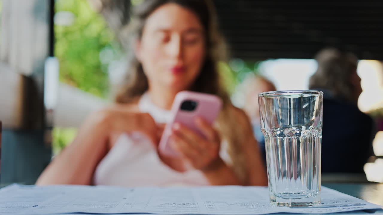 Close up of an empty glass on a table with a blurred view of a woman scrolling on her phone at a restaurant