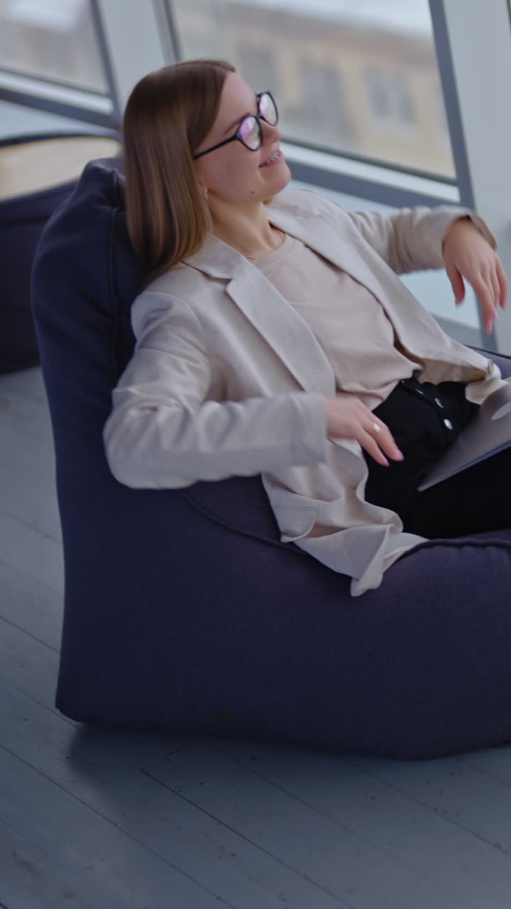 Happy relaxed woman sits in bag armchair with computer on her laps. Dreamy lady stretches hands and puts them behind her head. View from above. Vertical video