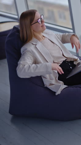 happy relaxed woman sits in bag armchair with computer on her laps. dreamy lady stretches hands and puts them behind her head. view from above. vertical video