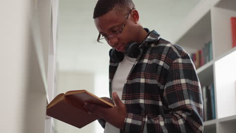 hipster reads book in college library. african american man with textbook learns information by bookshelf. passionate reader studies scientific literature