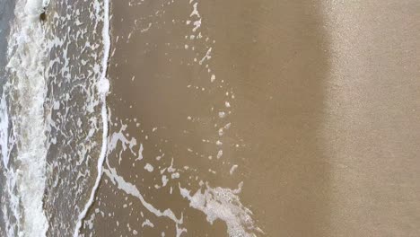 side-tracking-shot-of-the-beach-shore-with-sand-and-oals-of-the-sea,-vertical