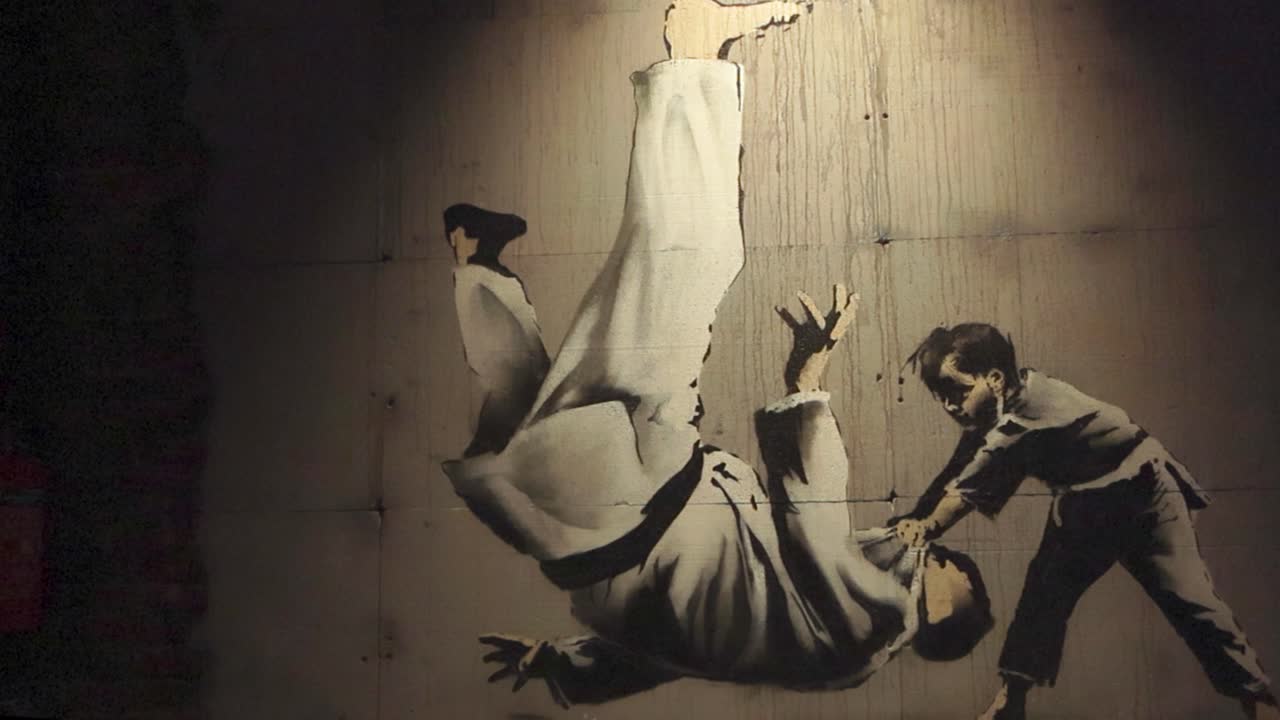 Two people performing martial arts or judo graffiti stencil art made by Banksy done on a concrete wall at his exhibition in Telliskivi. The artwork is well lit and shows a boy throwing an adult.