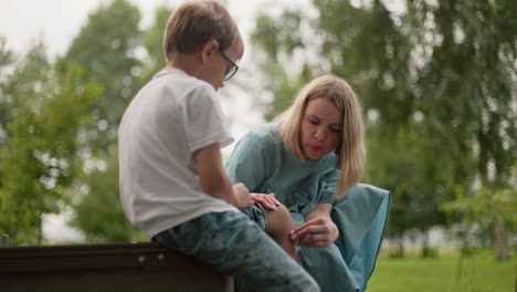 a woman in a blue dress blows gently on a boy's injured leg while cleaning it, showing care and tenderness