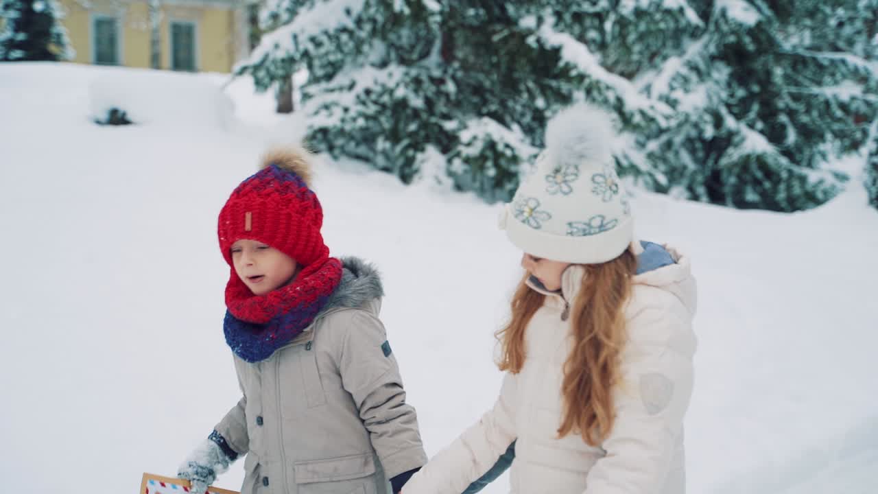 Cute girl in a white hat with flowers and a boy in a red hat are walking and looking at a sealed envelope in the middle of the road surrounded by fir-trees in the winter. Close-up. Slow motion
