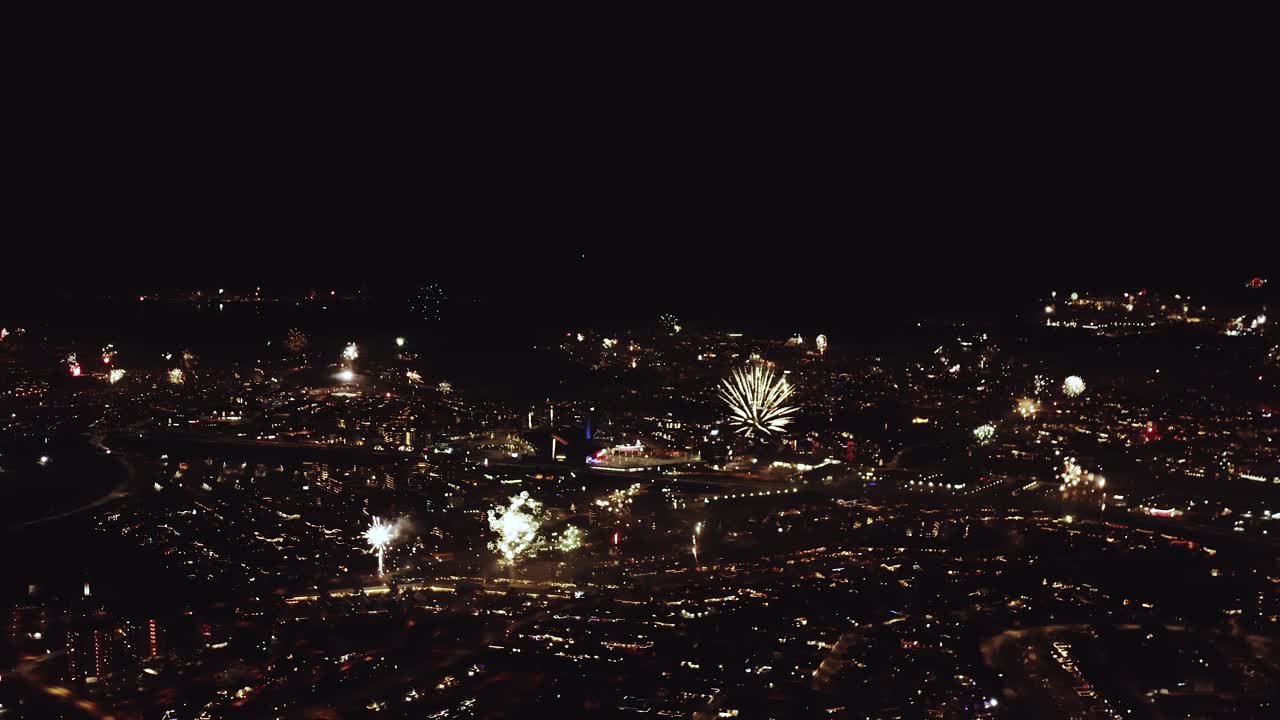 Ampia vista aerea di Reykjavík illuminata da fuochi d'artificio che esplodono sopra il paesaggio urbano. Strade, tetti e quartieri risplendono di luci festive, catturando una vivace atmosfera di festa