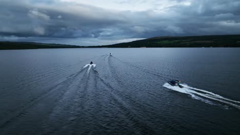 ángulo-Aéreo-De-Barcos-De-Alta-Velocidad-Que-Pasan-Por-Loch-Lomond,-Escocia-En-Una-Noche-Nublada