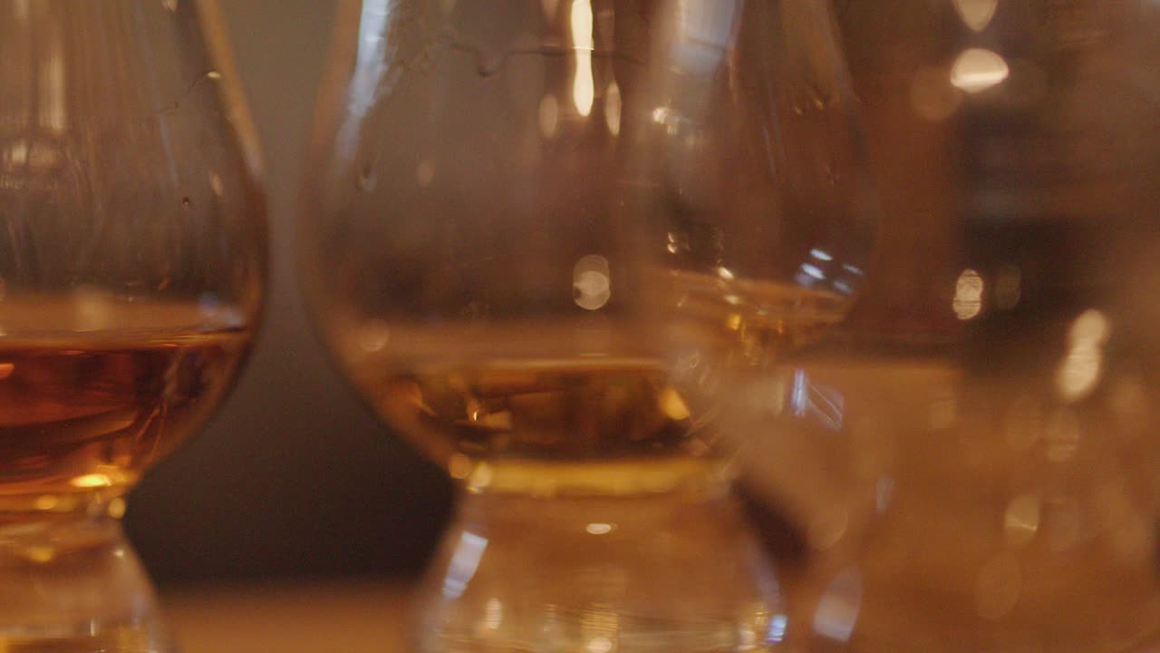 Multiple whiskey tasting glasses filled with amber liquid are shown in a warm, softly lit bar setting. Slow horizontal camera pan enhances depth