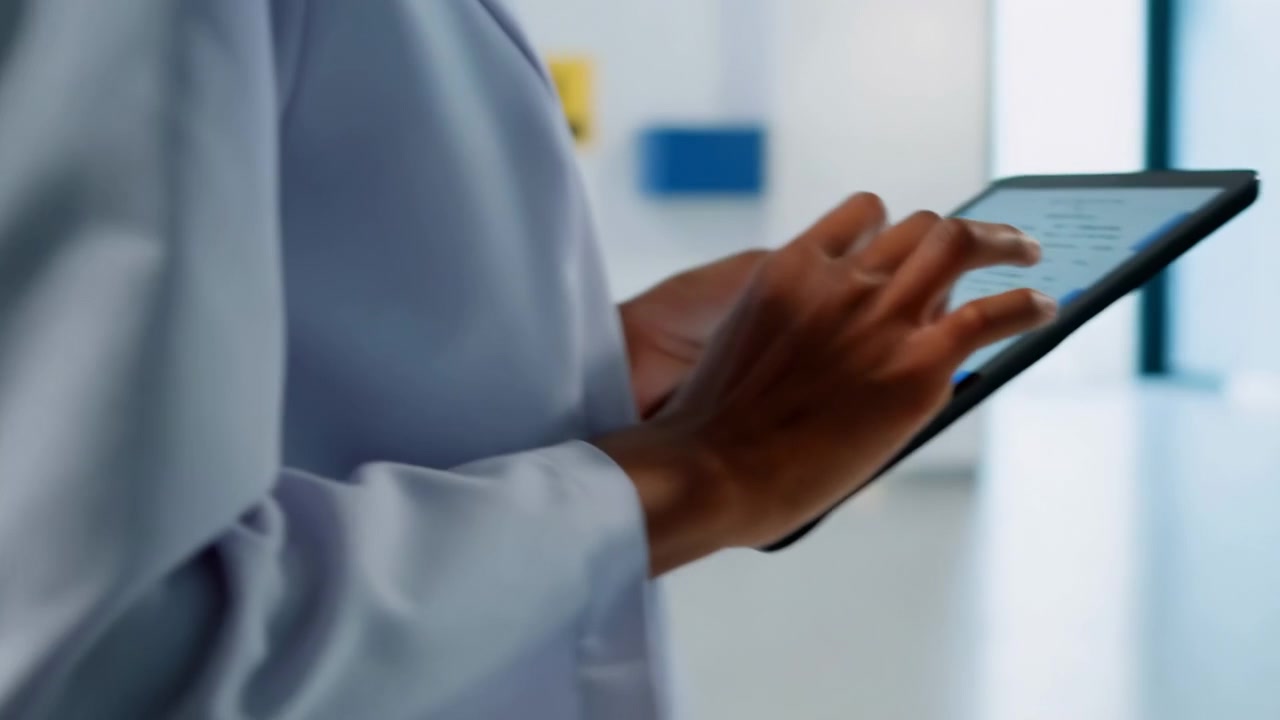 Close view of hands holding tablet in clinical corridor while fingertips tap, swipe, and review on-screen records, bright neutral light, clean surroundings, focused working posture, modern