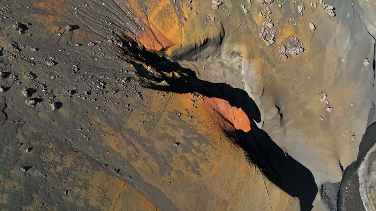 Top-down aerial close-up of a rugged volcanic slope with orange mineral streaks, dark shadowed ravines, and scattered rocks, revealing dramatic abstract geology and raw mountain texture.