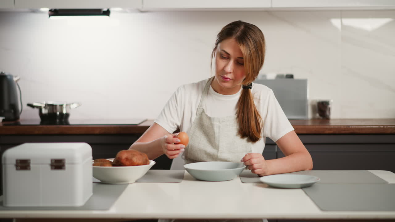 Focused female baker in white shirt and apron preparing to break egg on glass plate while seated at kitchen table, surrounded by potatoes, utensils, and appliances in clean domestic cooking area