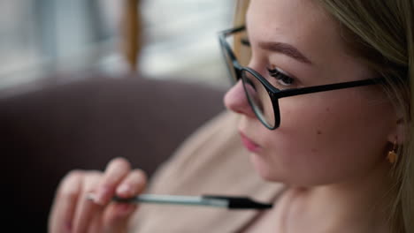 primer plano de una mujer blanca pensativa con la pluma suavemente presionada contra los labios, tocando pensativamente contra ellos mientras lleva gafas, fondo borroso que muestra contemplación o pensamiento profundo