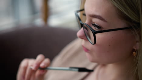 close-up of pensive white lady with pen softly pressed against lips, thoughtfully tapping against them while wearing glasses, blurred background showcasing contemplation or deep thought