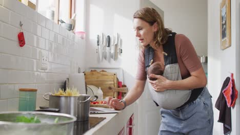 Video-of-caucasian-mother-with-newborn-baby-in-baby-carrier-using-laptop-in-kitchen