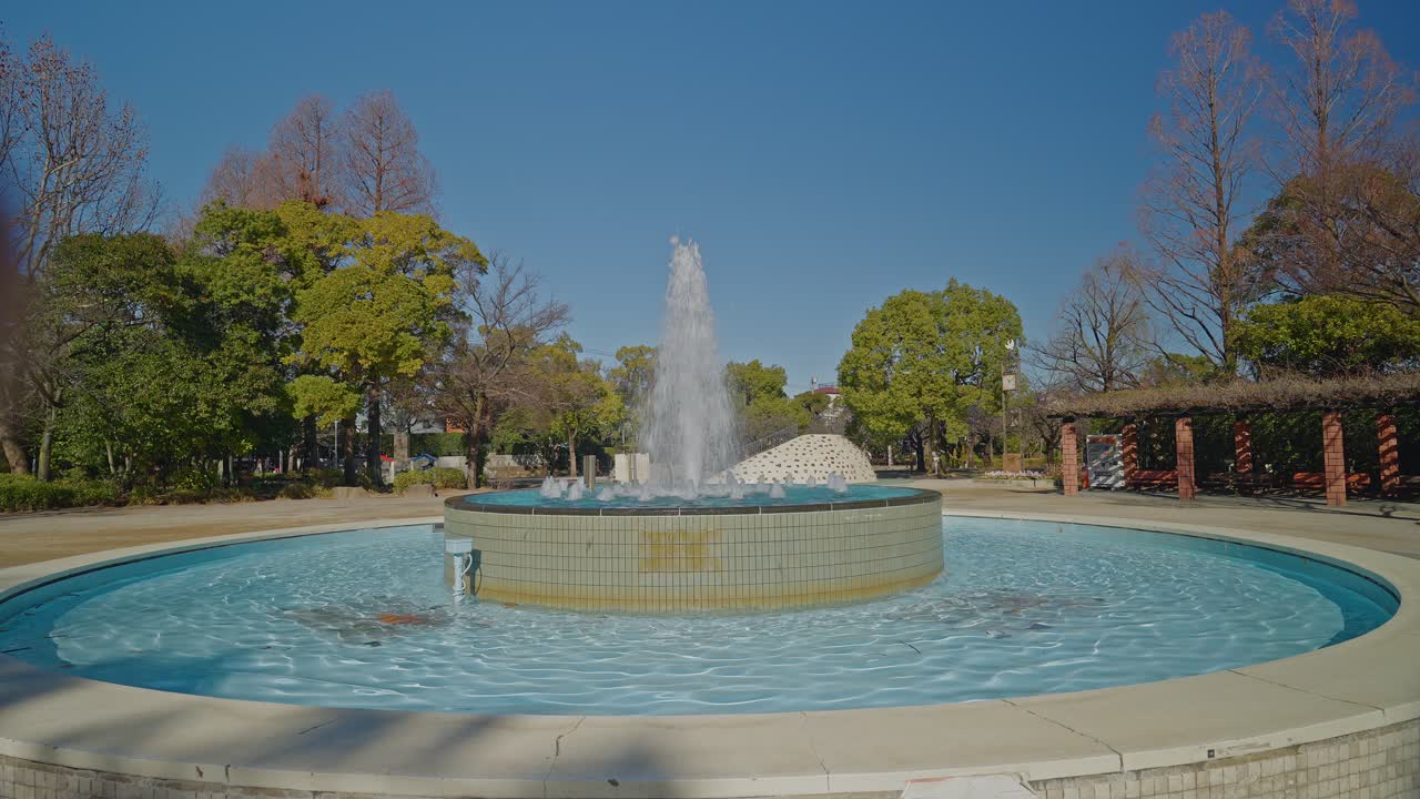 Un plano general de una fuente de agua pulverizada en el centro del Parque Gyosen, rodeada de árboles verdes bajo un cielo azul despejado