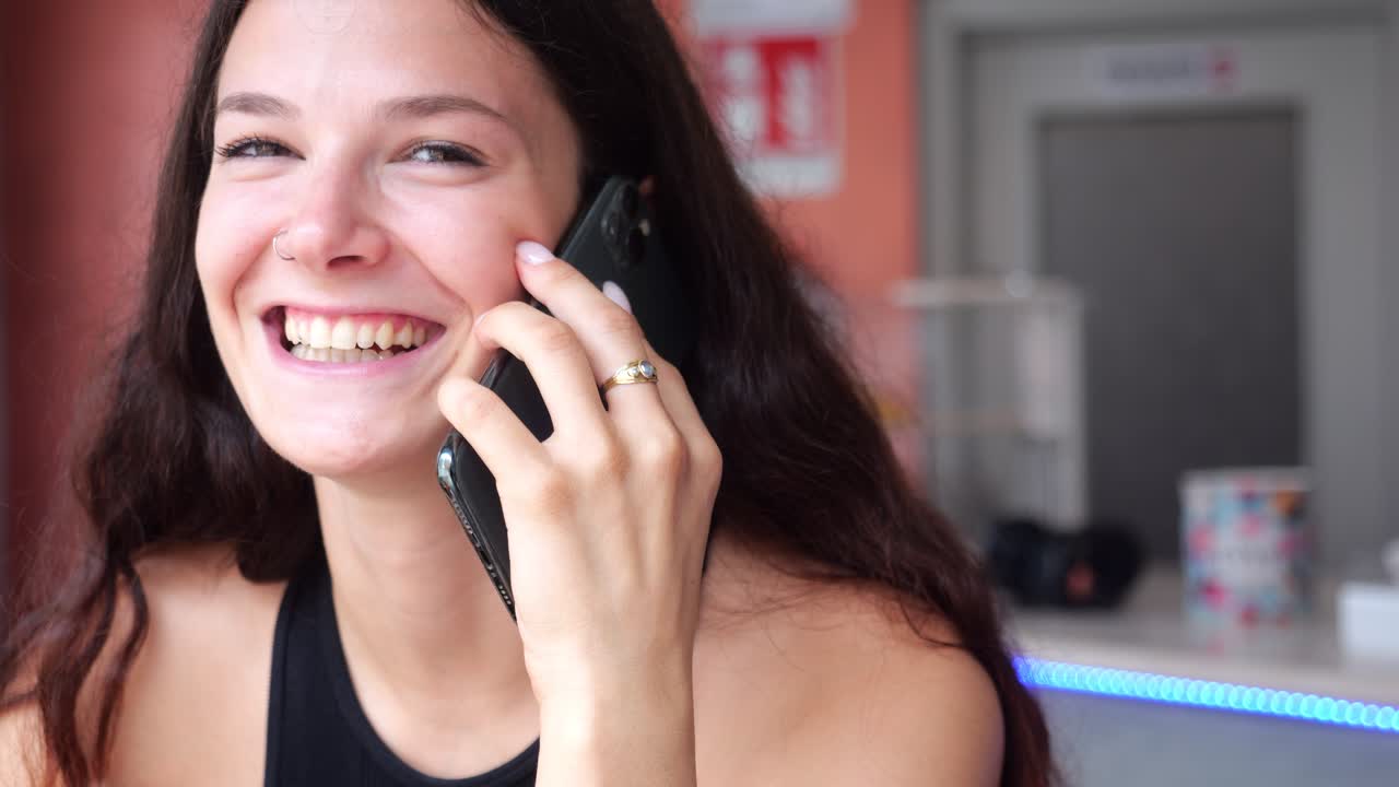 Woman with dark hair holds smartphone to ear in relaxed bar environment, conversing casually against warm orange-toned interior with soft ambient lighting and blurred background elements