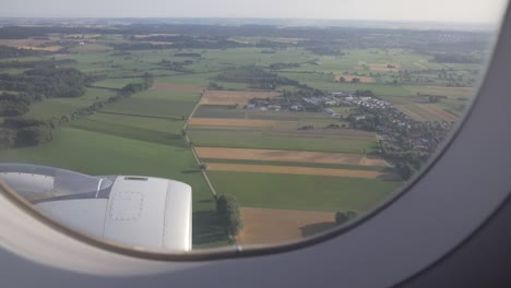 green fields, trees and european settlement are seen through window of aircraft.